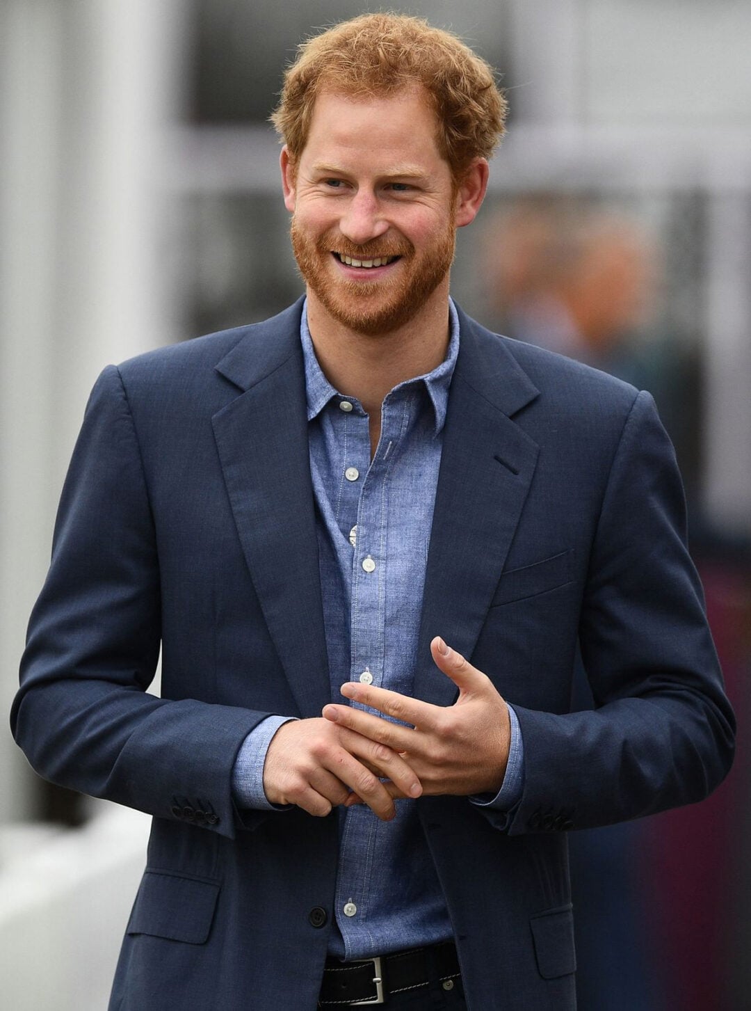 A man with reddish hair and a beard smiles while wearing a blue suit jacket over a light blue button-up shirt. He stands outdoors with his hands loosely clasped.