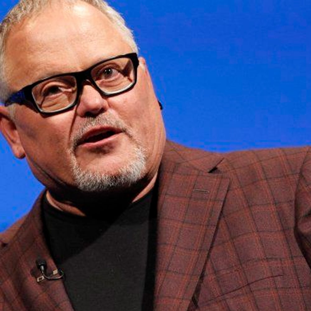A man with short light hair, glasses, and a goatee wears a brown plaid jacket over a black shirt. He appears to be speaking or presenting, with a microphone clipped to his jacket.