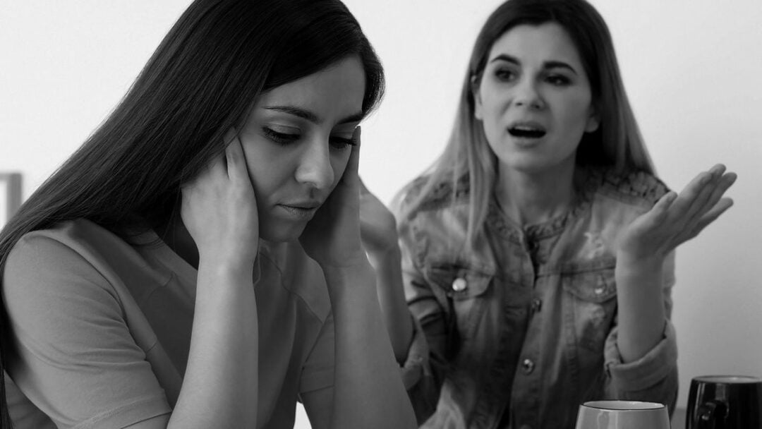 A young woman sits with her hands covering her ears, looking upset, while another woman beside her gestures and speaks passionately.