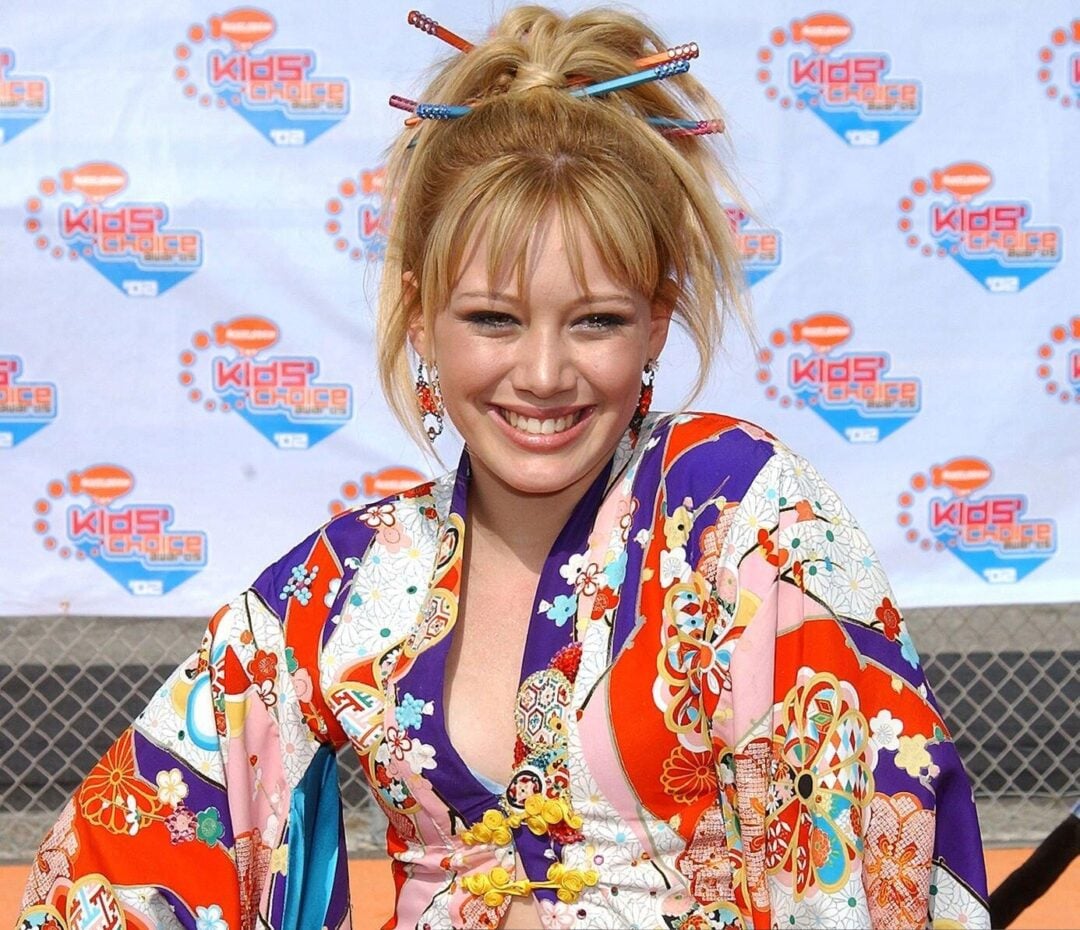 A young woman smiling on a red carpet, wearing a colorful, floral kimono over a purple top and jeans. Her hair is styled up with chopsticks, and she stands confidently with her hands on her hips.