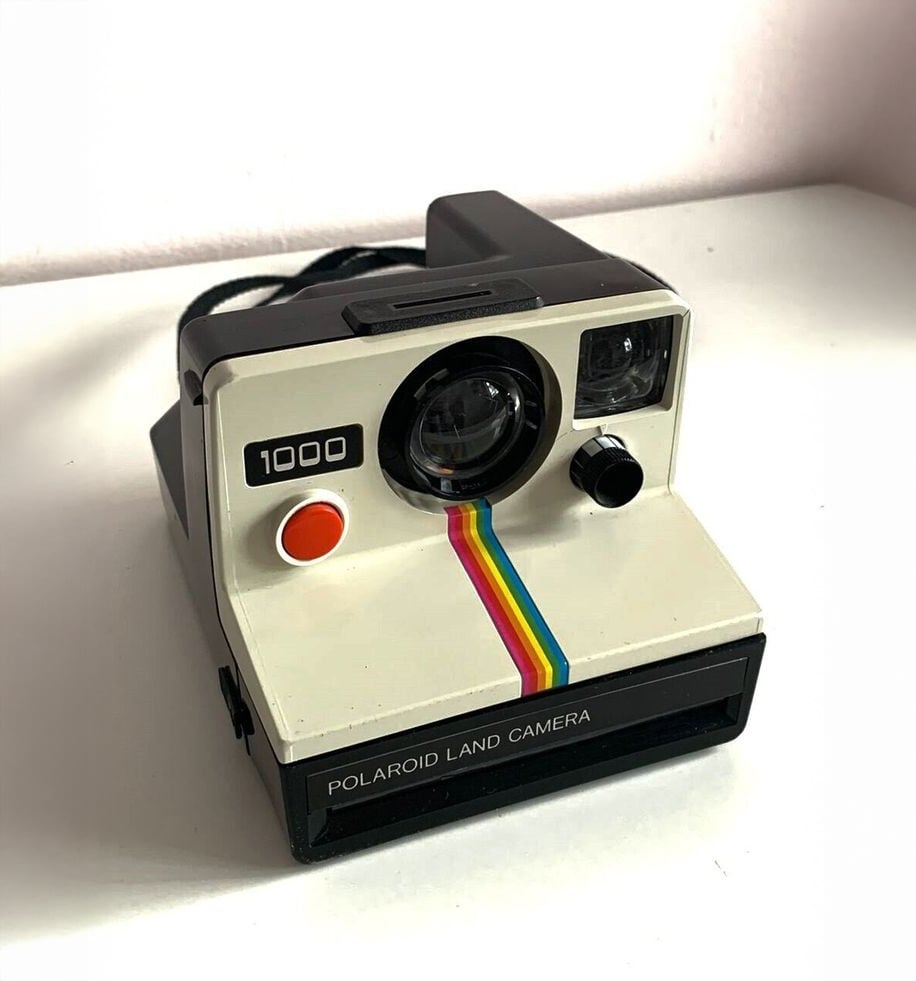 A vintage Polaroid Land Camera 1000 with a rainbow stripe on the front sits on a white surface against a light-colored wall.