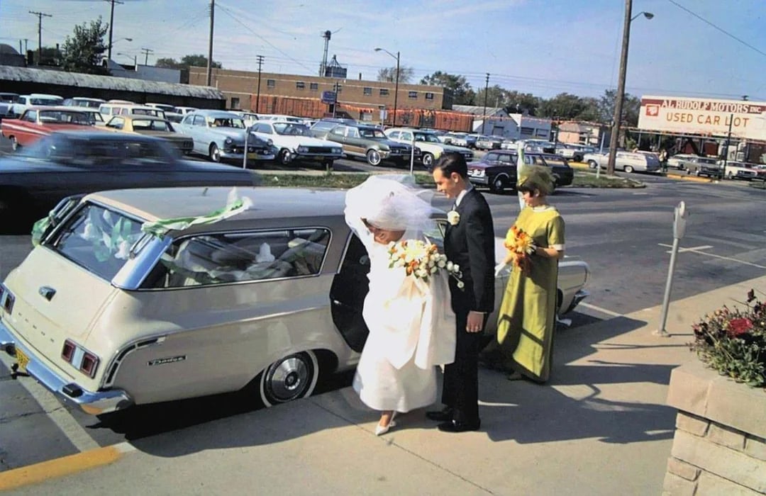 A bride in a white dress and veil steps into a vintage white station wagon, assisted by a man in a suit, while a woman in a green dress holding flowers stands nearby. Classic cars and a used car lot are visible in the background.
