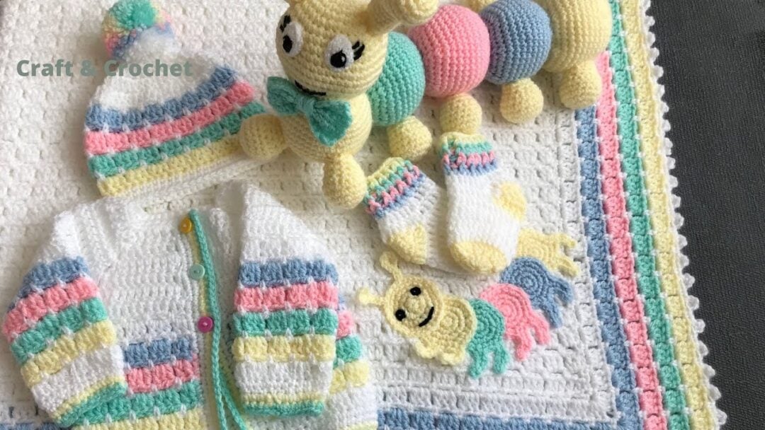 Crocheted baby items including a striped cardigan, matching hat, socks, an amigurumi caterpillar, and a blanket with colorful borders. The set features soft pastel shades of yellow, pink, blue, and green.
