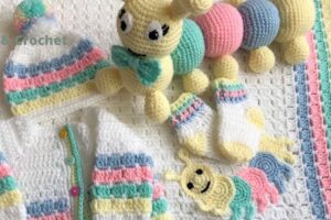 Crocheted baby items in white, pastel pink, blue, yellow, and green include a cardigan, hat with pom-pom, socks, a striped blanket, a large caterpillar toy, and a small caterpillar applique.