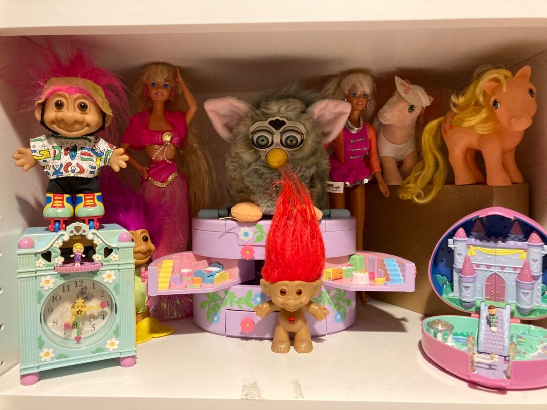 A shelf displays a variety of vintage toys, including a troll doll, Barbies, a Furby, My Little Pony figures, a pastel toy clock, and a small plastic castle playset with bright colors and playful designs.
