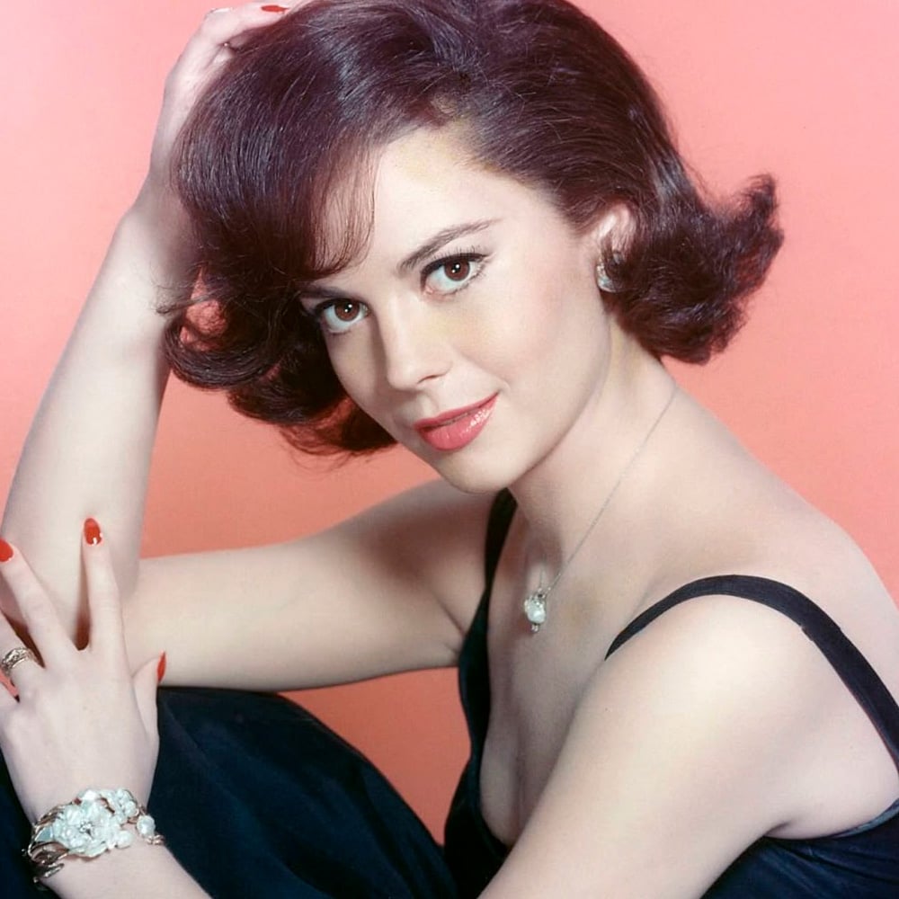 A woman with short, wavy brown hair poses against a pink background. She wears a black dress, red lipstick, and red nail polish, accessorized with earrings, a bracelet, and a necklace, looking at the camera.