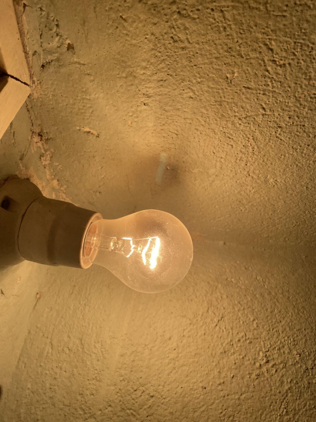 A lit incandescent light bulb is mounted on a textured, beige wall, casting a warm glow and creating soft shadows around it.