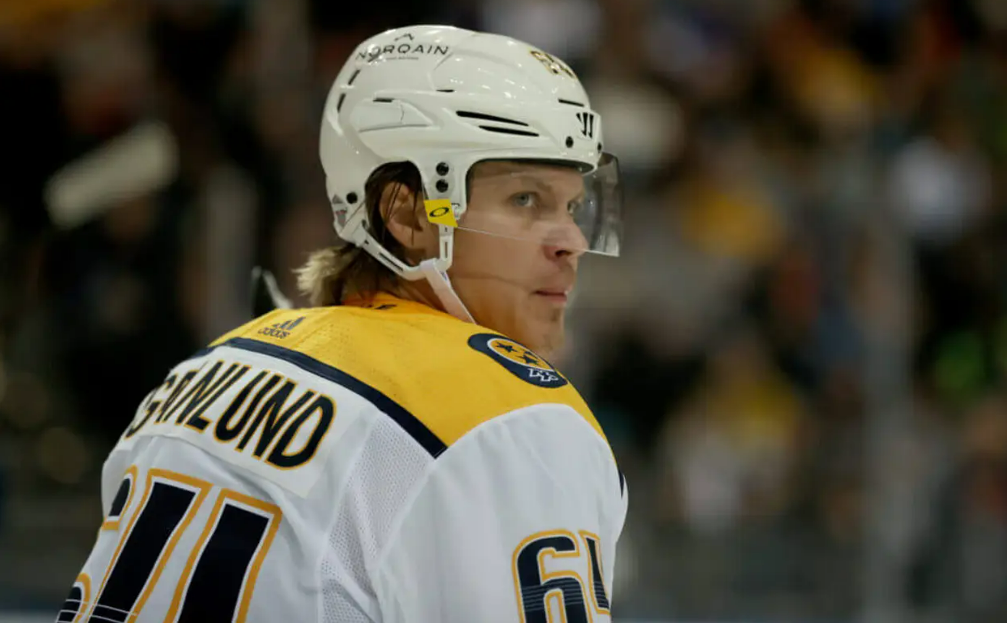 A hockey player wearing a white and yellow jersey with the name “GRANLUND” and number 64 on the back looks over his shoulder during a game, with the blurred crowd in the background.