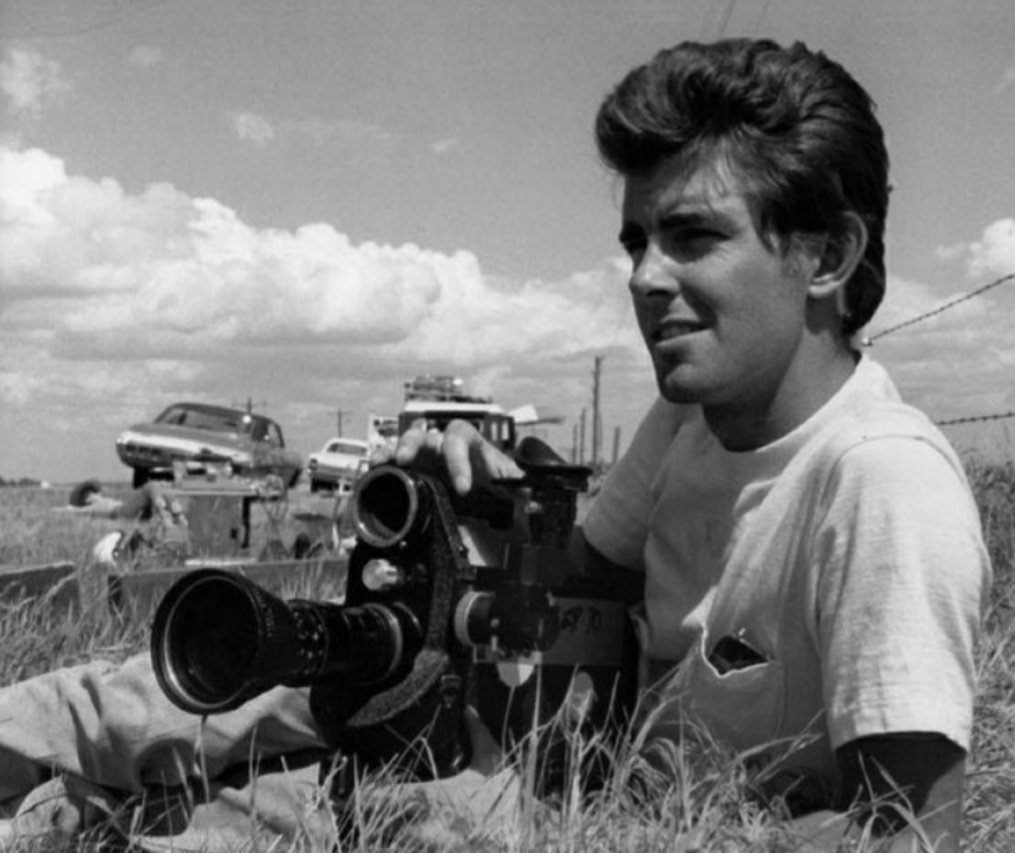 A young man with tousled hair lies in tall grass, holding a vintage film camera. Old cars are parked on a dirt road behind him under a partly cloudy sky.