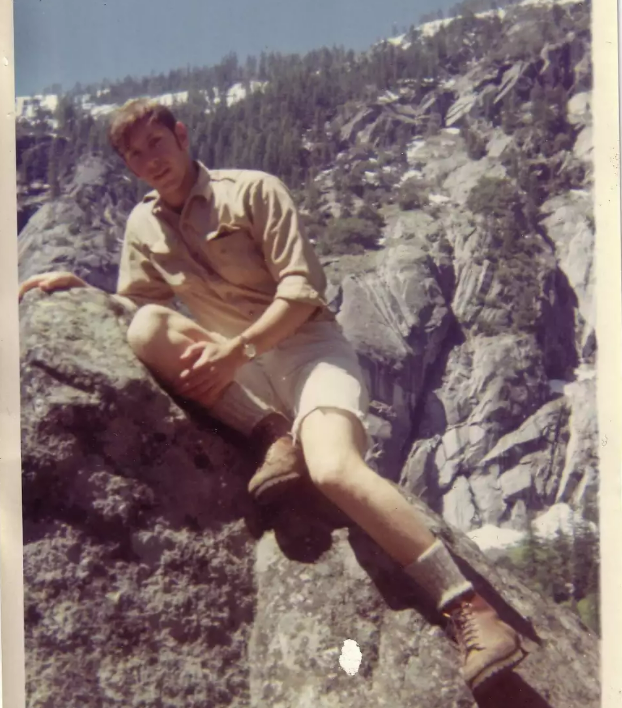 A young man in a tan shirt and shorts sits on a large rock with one leg extended, wearing hiking boots and socks. Rocky cliffs and pine trees are visible in the background under a clear blue sky.