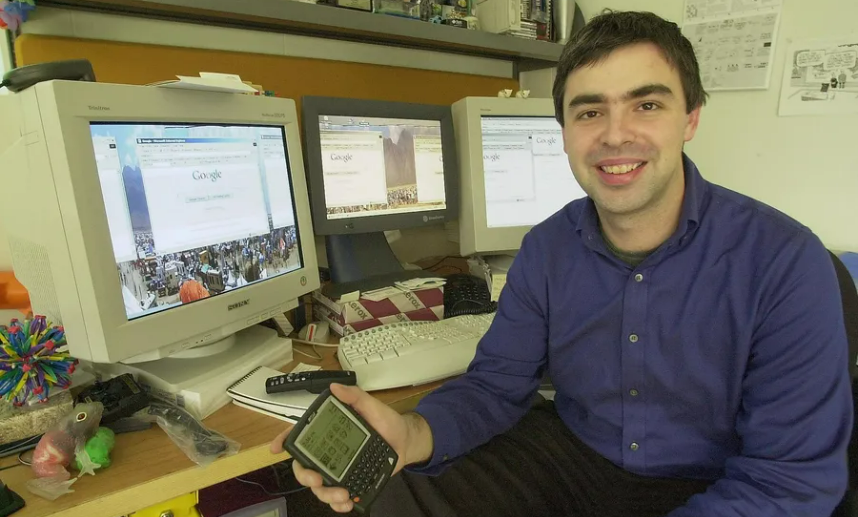 A man in a blue shirt sits at a cluttered desk with three old computer monitors displaying Google’s homepage. He is smiling and holding an early handheld device. The workspace has various office items and computer accessories.