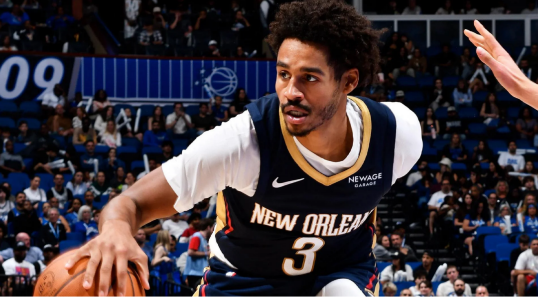 A basketball player wearing a New Orleans jersey, number 3, dribbles the ball while being guarded during a game, with a crowd and blue court background.