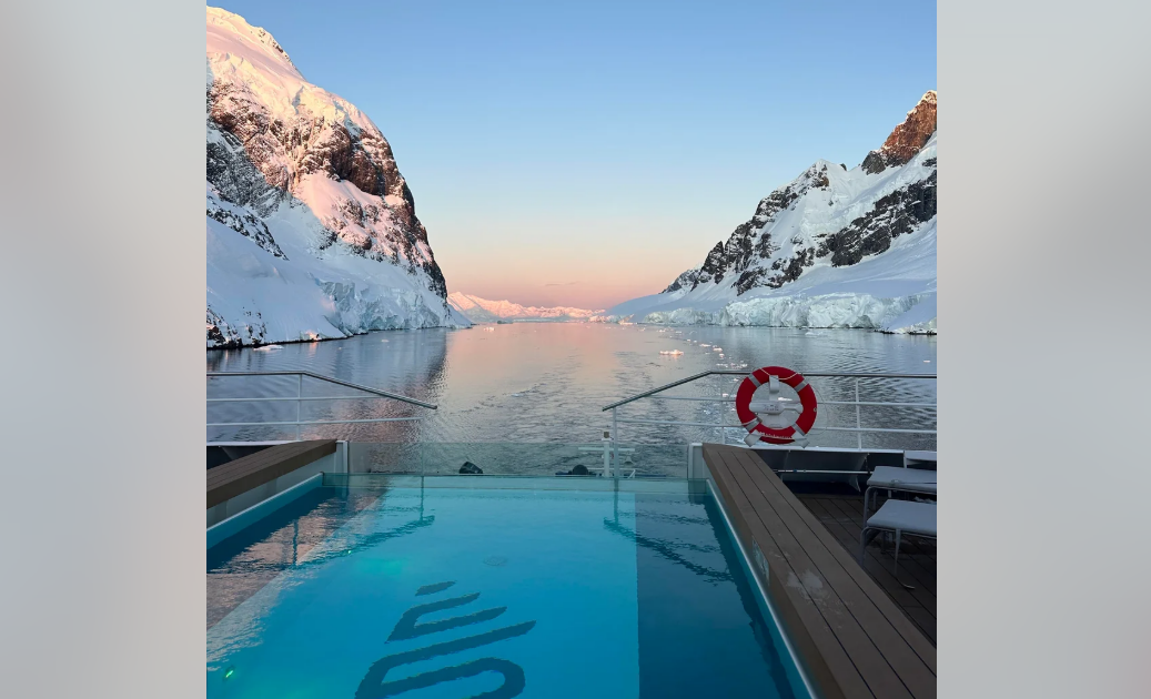 A swimming pool on a ship overlooks calm icy waters, flanked by snow-covered mountains at sunset, casting a soft pink and blue glow on the landscape. A red lifebuoy hangs on the railing to the right.