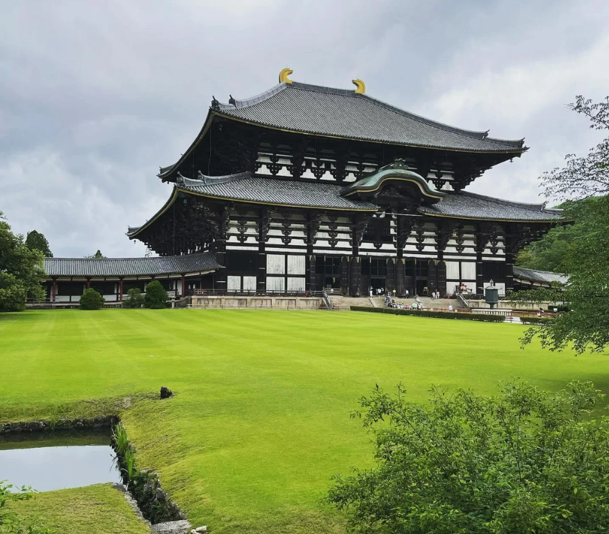 Large traditional Japanese wooden temple with a curved roof and ornate details, surrounded by a green lawn and trees under a cloudy sky.