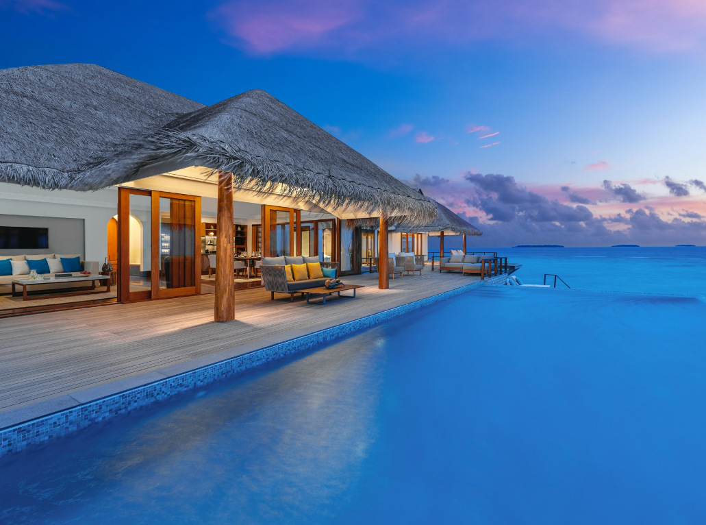 A luxurious beachfront villa with a thatched roof, large glass doors, cozy outdoor seating, and an infinity pool overlooking a calm, blue ocean at sunset. The sky is tinged with orange, pink, and purple hues.