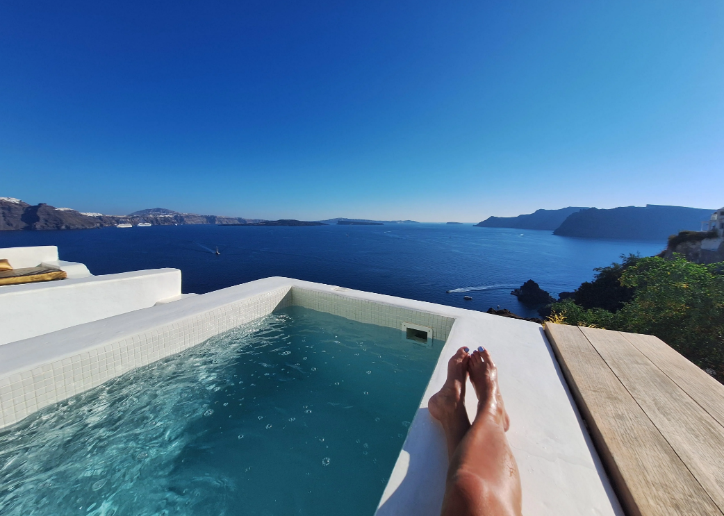 Person relaxing with their legs in a small pool, overlooking a stunning blue sea and distant cliffs under a clear sky, with modern white architecture on the left and greenery to the right.