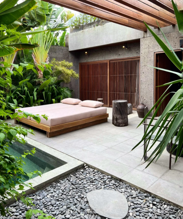 An outdoor bedroom with a minimalist wooden bed, pink bedding, and two pillows, surrounded by lush green plants, a small pool, a stone path, and wooden lattice doors under a pergola.