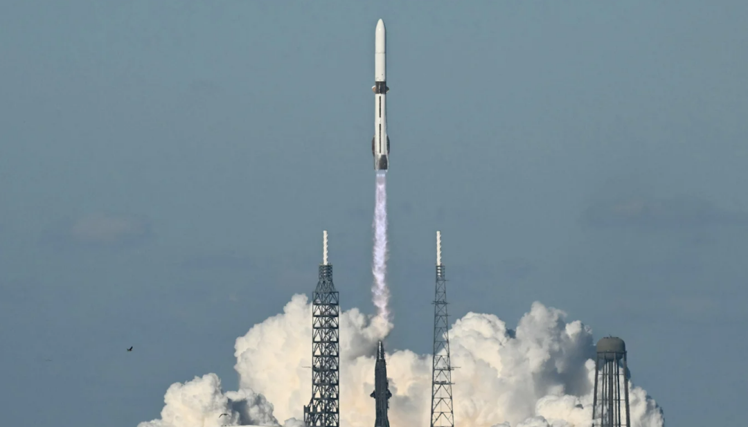 A rocket launches into the sky, leaving a trail of smoke and flames, with clouds billowing at the base and support structures visible on either side. The sky is clear and blue in the background.