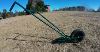A green metal wheel hoe with a single large black wheel rests on dry grass in an open, rural field with trees and a wooden structure in the background under a clear blue sky.