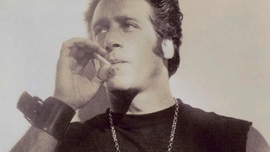 A man with styled dark hair smokes a cigarette while wearing a sleeveless shirt, a large wrist cuff, and a chain necklace. The photo is in black and white, giving it a vintage or retro look.