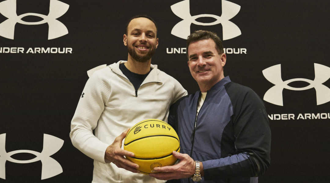 Two men stand in front of a backdrop with Under Armour logos. One holds a yellow basketball labeled "CURRY." Both are smiling and wearing casual jackets.