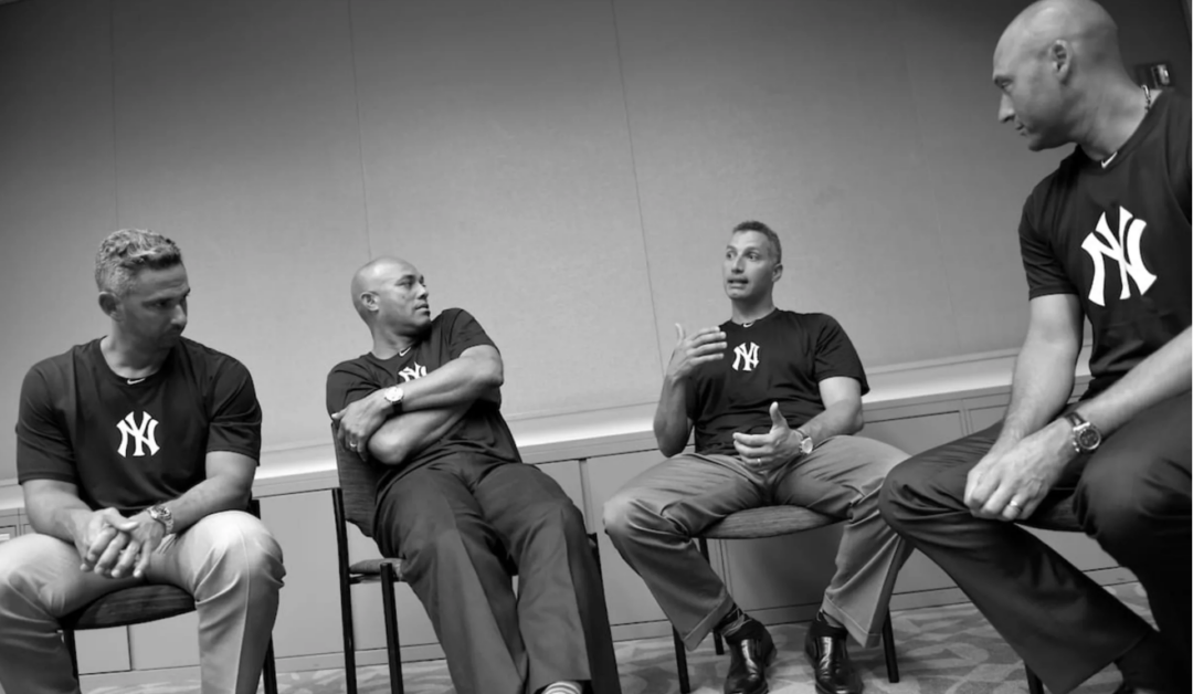 Four men wearing New York Yankees shirts sit in chairs in a plain room, engaged in conversation. The image is black and white, and the men appear relaxed and attentive.