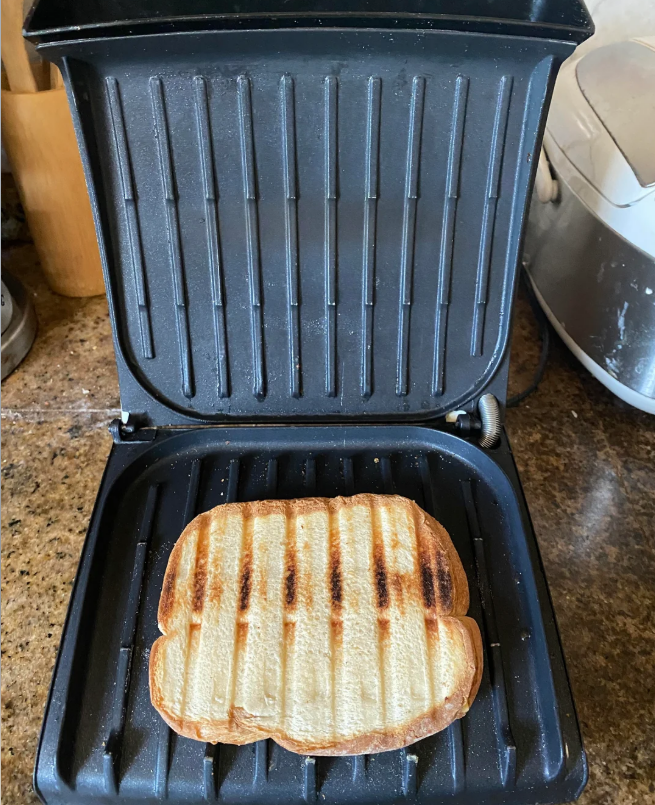 A toasted sandwich sits on the grill of an open sandwich press on a kitchen countertop. The bread has visible grill marks and slight charring. Kitchen appliances and utensils are in the background.