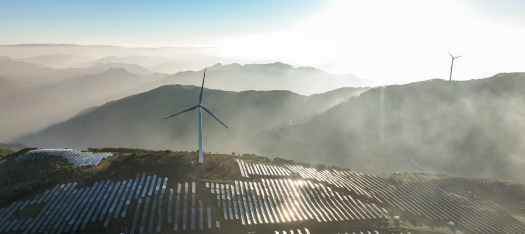 A wind turbine stands among rows of solar panels on a mountain ridge, with mist and sunlight illuminating the landscape and distant hills in the background.