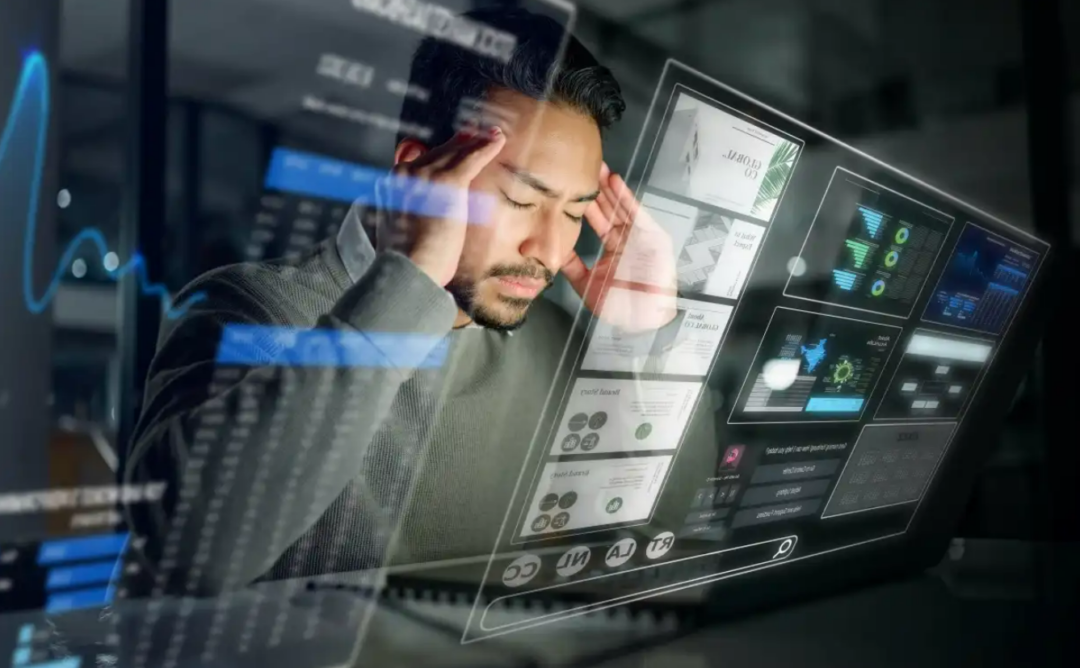 A man holding his head in frustration sits in front of a futuristic transparent screen displaying charts, data, and digital interfaces in a dark office setting.