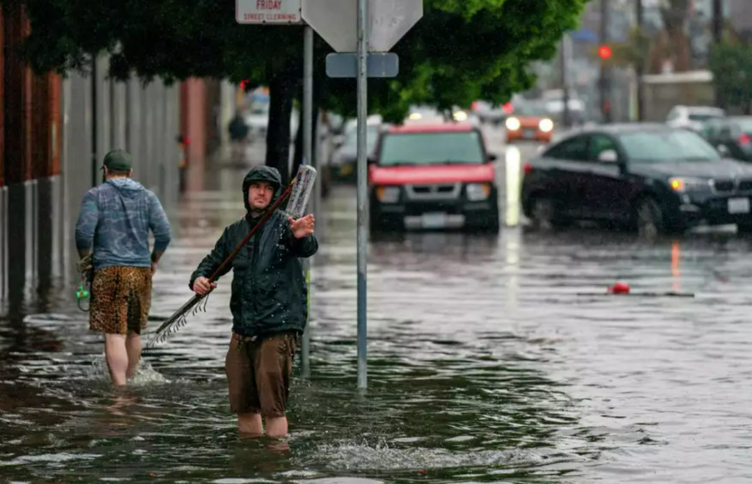 Two people walk through a flooded city street. One person faces the camera holding a long object over their shoulder, while the other walks away. Parked cars are partially submerged in water in the background.