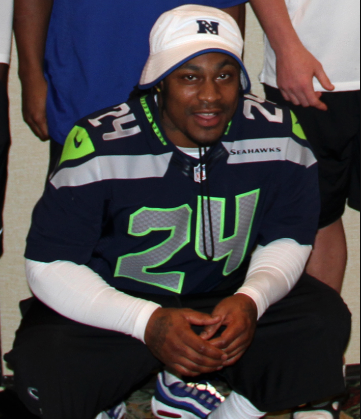 A man wearing a white bucket hat and a Seattle Seahawks football jersey with the number 24 squats and smiles at the camera.