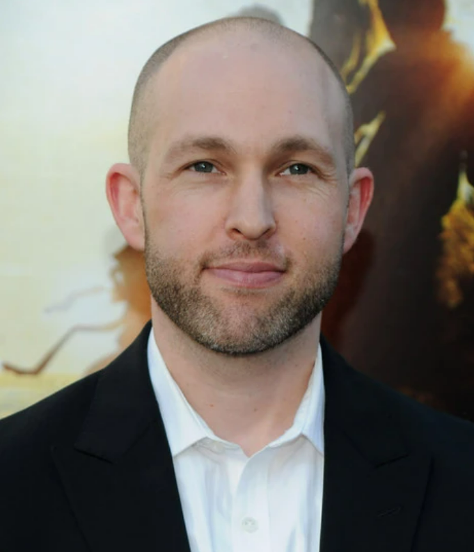 A man with a bald head and trimmed beard, wearing a white shirt and black jacket, stands in front of a blurred, warmly lit background.
