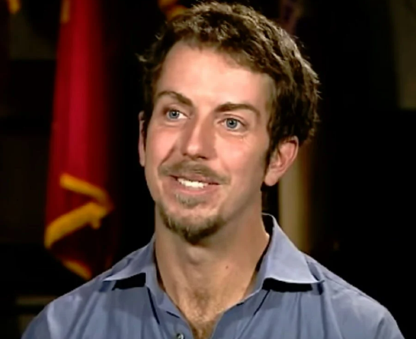 A man with short brown hair, light facial hair, and blue eyes is smiling. He is wearing a light blue collared shirt and sitting indoors with a blurred background featuring a red flag.