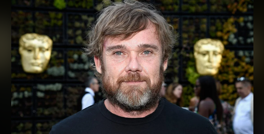 A man with light brown hair and a beard stands outdoors, wearing a black shirt. Behind him, two large gold masks are visible on a wall, and several people are blurred in the background.