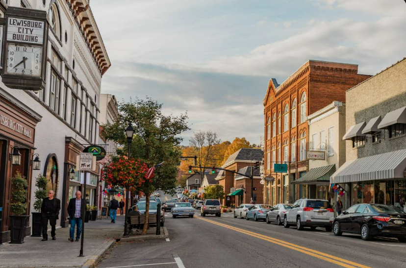A small town main street with historic brick buildings, shops, parked cars, and people walking on the sidewalks under a partly cloudy sky. Trees with autumn foliage are visible in the background.