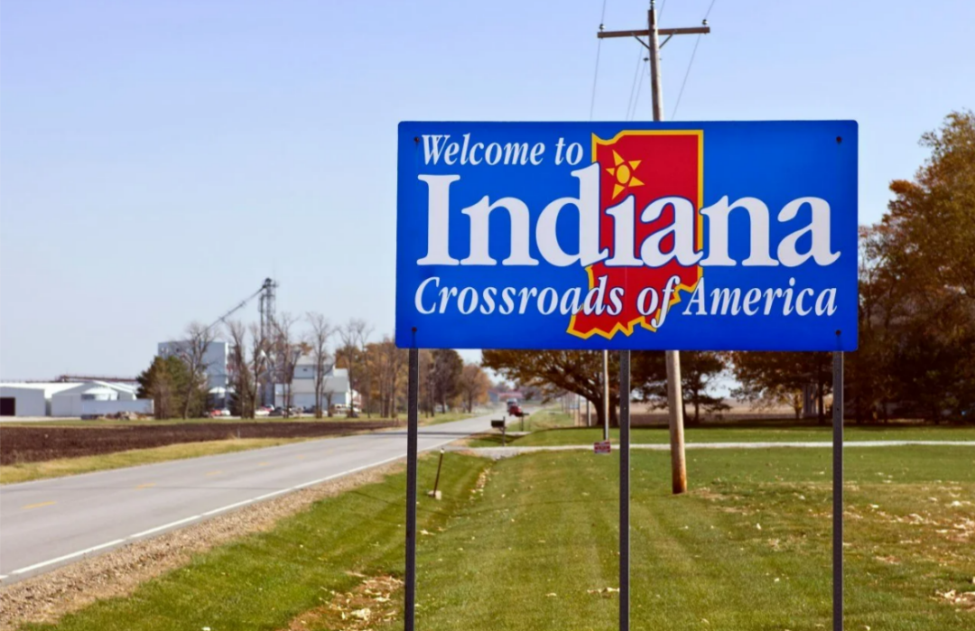 A blue roadside sign reads "Welcome to Indiana, Crossroads of America" with a yellow outline of Indiana’s state shape. The sign is next to a rural road with grass, trees, and some farm buildings.