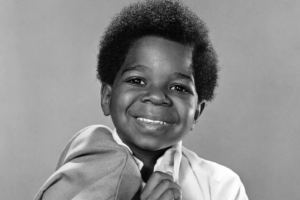 A young Black boy with a short afro smiles at the camera, holding a jacket over his shoulder. The photo is black and white with a plain background.