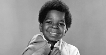 A young Black boy with a short afro smiles at the camera, holding a jacket over his shoulder. The photo is black and white with a plain background.