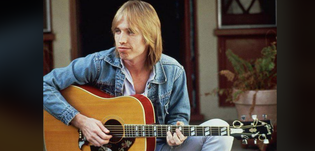 A man with straight, shoulder-length blond hair wearing a denim jacket plays an acoustic guitar while sitting outdoors in front of a building.