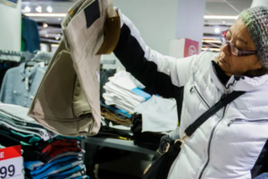A woman wearing glasses, a knit hat, and a white winter coat inspects a pair of beige pants in a clothing store. A sale sign shows a price of $14.99 near folded clothes on a table.