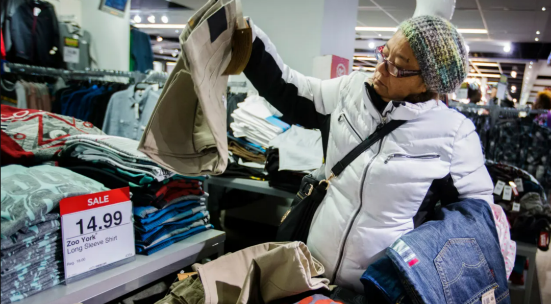 A woman in a white jacket and knit hat shops for pants in a clothing store, holding up a pair to inspect. A sale sign nearby reads "14.99 Zoo York Long Sleeve Shirt." Stacks of folded clothes fill the table.