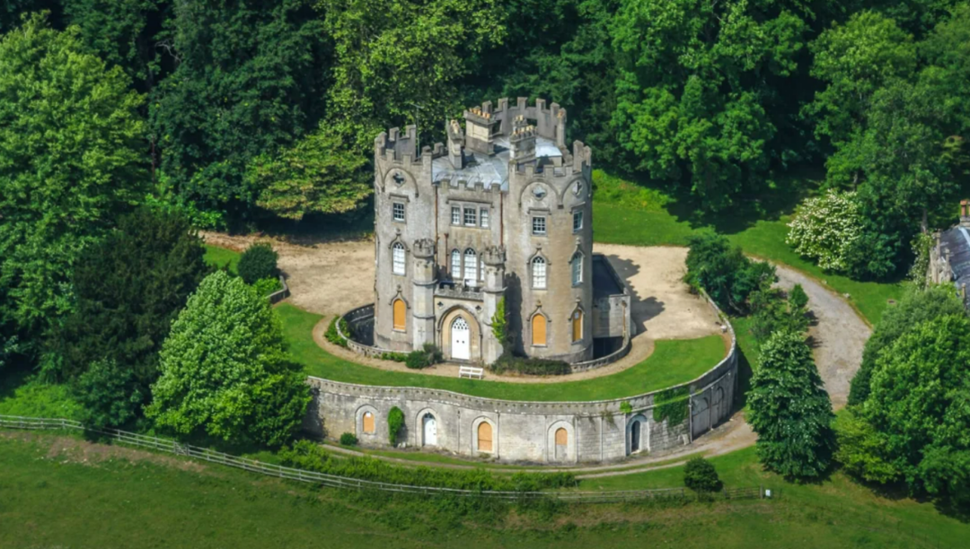 Aerial view of a small, round medieval-style stone castle with crenellated towers, surrounded by lush green trees and a circular stone wall, located in a grassy clearing.
