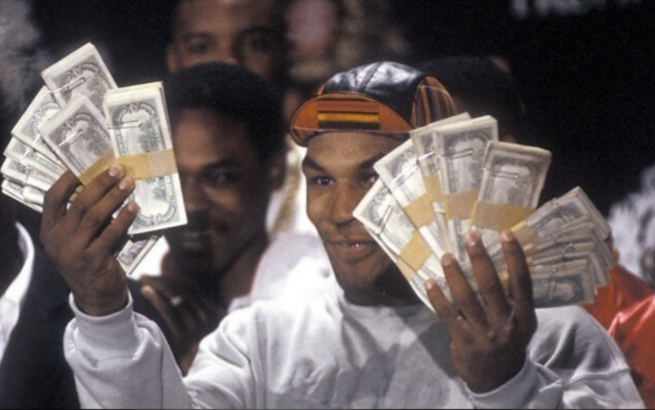 A man smiling and holding up large stacks of cash in both hands, surrounded by people in the background.