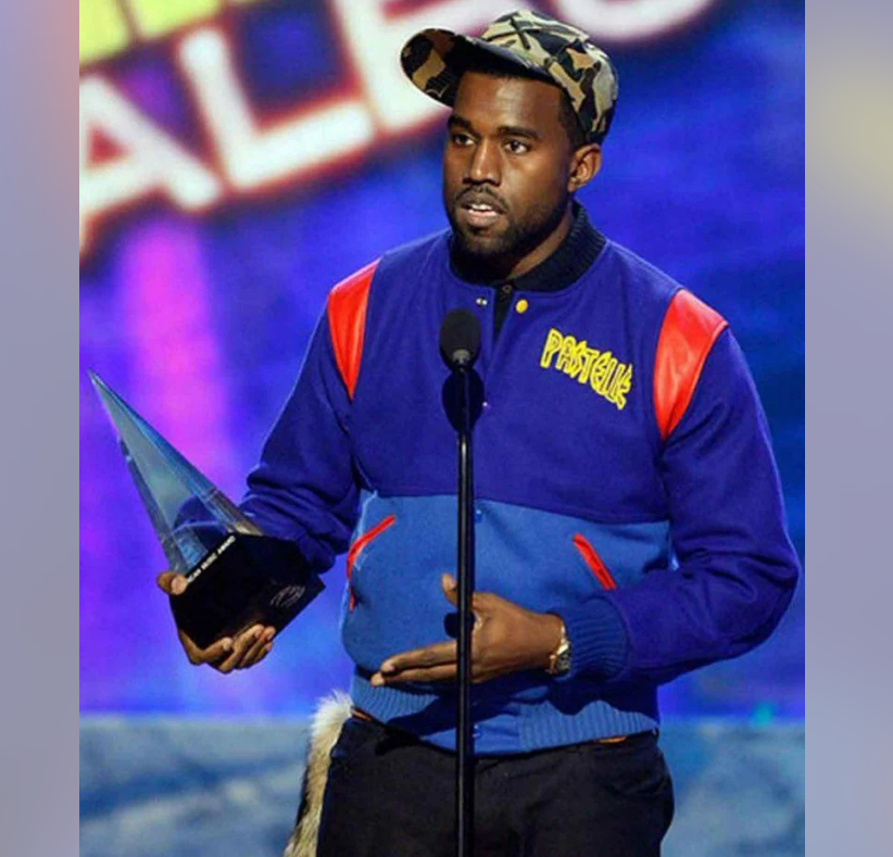A man wearing a colorful jacket and a camouflage cap holds a triangular award trophy while speaking into a microphone on stage, against a blurred blue and purple background.