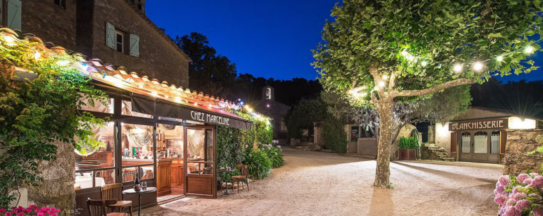 A charming village scene at dusk with a warmly lit café, “Chez Marguerite,” outdoor seating, string lights in trees, and a bakery labeled “Boulangerie” in the background.