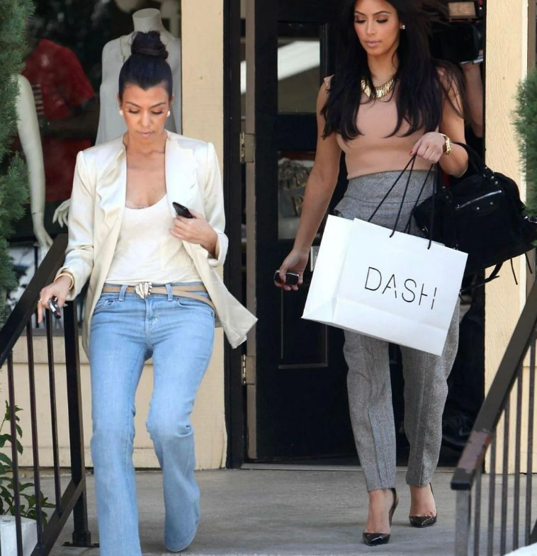 Two women walk down steps outside a building. One wears light blue jeans, a beige top, and a white blazer; the other wears grey pants, a tan top, and carries a white shopping bag labeled “DASH.”