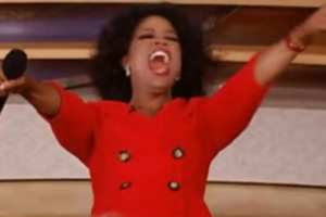 A woman in a bright red outfit stands with arms outstretched, enthusiastically shouting while holding a microphone. She appears excited and joyful on a stage or set.