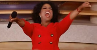 A woman in a bright red outfit stands with arms outstretched, enthusiastically shouting while holding a microphone. She appears excited and joyful on a stage or set.