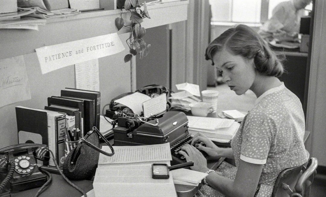 A woman in a polka-dot dress types on a typewriter at a cluttered desk with papers, books, and a rotary phone. A sign above her reads "Patience and Fortitude." Another person works in the background.