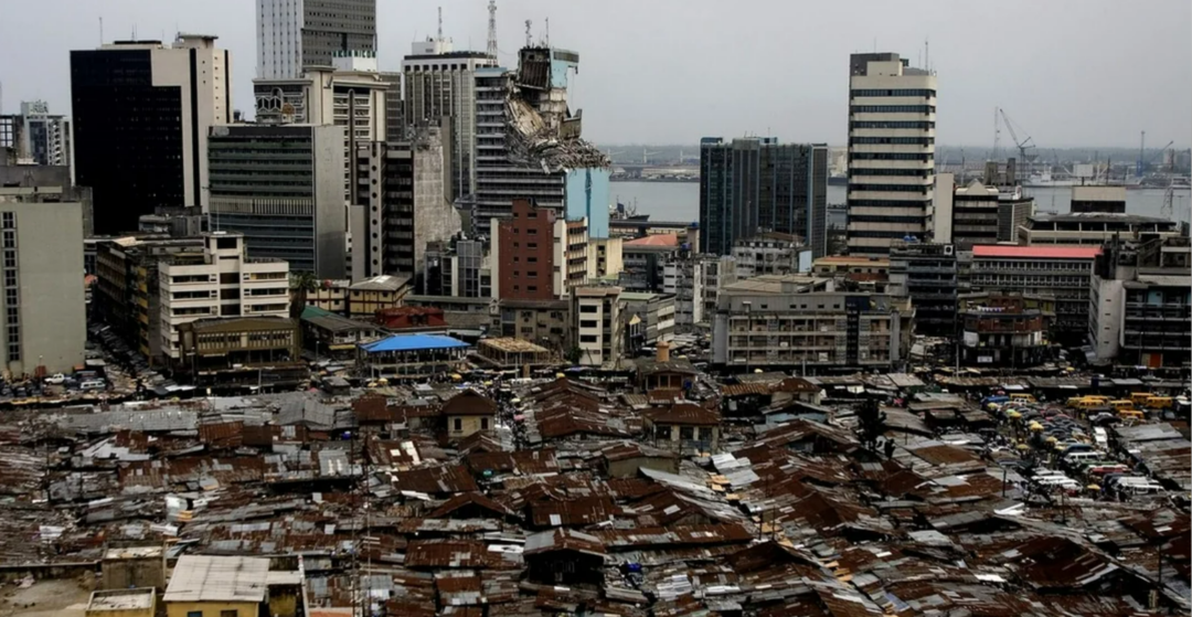 A cityscape showing a stark contrast between densely packed, rusted-roof slums in the foreground and modern high-rise buildings in the background under a cloudy sky.